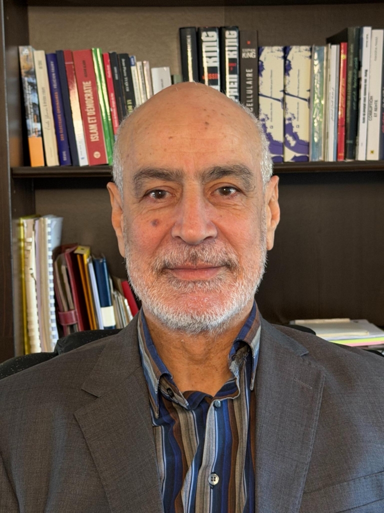 Photo of Fouad Abdelmoumni who is in front of a bookcase filled with books. He has facial haird, and wears a striped shirt and blazer.