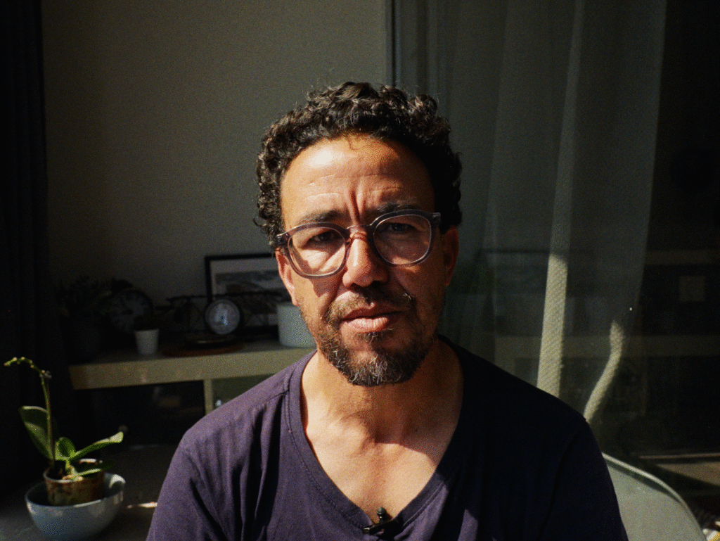 Photo of Hicham Mansouri facing the camera. He has curly hair and facial hair, and is wearing glasses and a t-shirt.